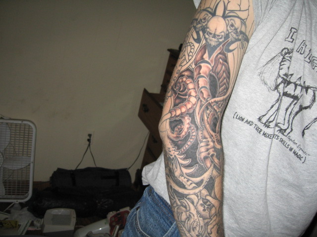 Man's forearm with black-and-gray biomechanical sleeve tattoo, skull at top, indoor room