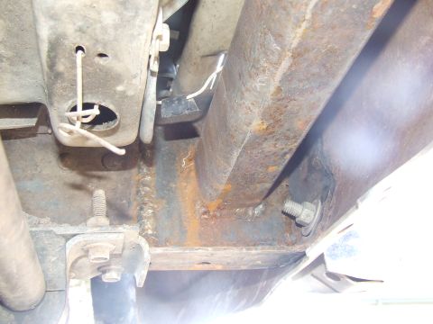 1986 Ford Bronco - Underbody close-up of a rusty frame rail and crossmember area showing a welded seam, a bolted bracket with an exposed threaded stud, a whit…