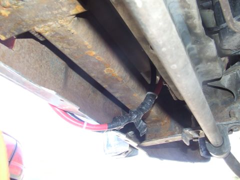 1986 Ford Bronco - Underside close-up of a surface-rusted boxed frame rail with heavy-gauge red and black battery cables routed along it and secured with tape…