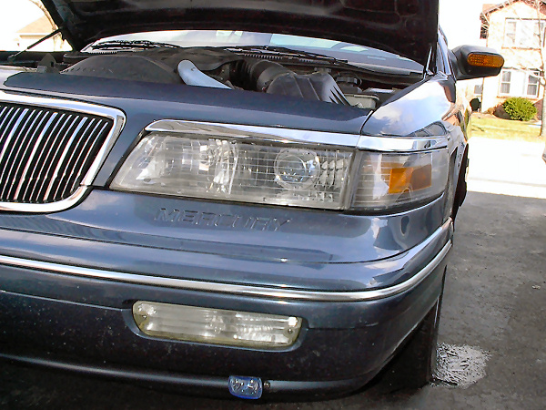 Front corner of blue-grey Mercury sedan with hood up, chrome headlight trim and fog lamp