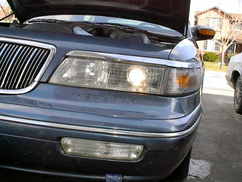 1985 Mercury Grand Marquis - The front of a dark blue Mercury Grand Marquis is visible with the hood up, revealing the engine bay and chrome grille.