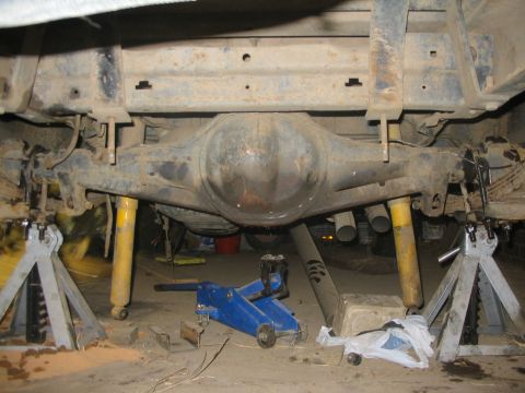 1981 Ford Bronco - Underside view of a vehicle's rear axle and suspension components supported by jack stands, with a blue hydraulic jack nearby.