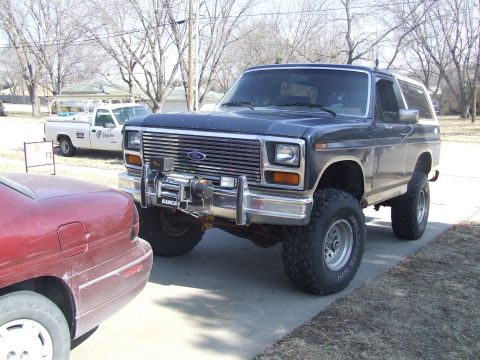 1986 Ford Bronco - A dark blue Ford Bronco is parked on a driveway next to a red car, with a white pickup truck in the background.