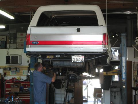 1990 Ford Bronco - white Ford Bronco is raised on a two-post lift from the rear as a mechanic works on the undercarriage.