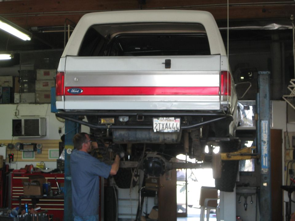 White Ford Bronco raised on a shop lift from the rear while a mechanic works underneath