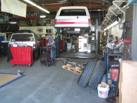 1990 Ford Bronco - Interior of a 4WD repair shop with a white Bronco on a lift, tool carts, a removed axle and stacked off-road tires.
