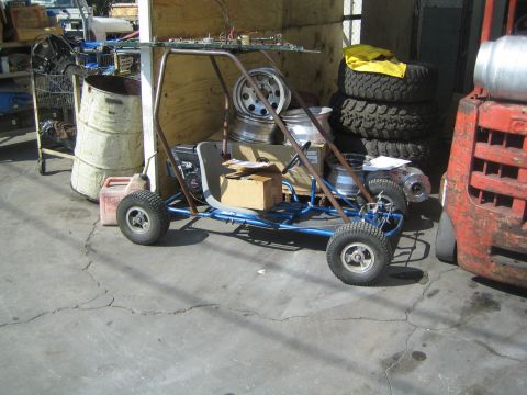 1990 Ford Bronco - homemade blue-tube-framed go-kart sits on a concrete shop floor against plywood, beside a barrel and stacked tires.