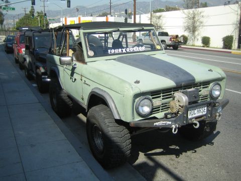 1990 Ford Bronco - light green early Ford Bronco with a front winch, black hood stripe and the top removed parked curbside on a sunny street.