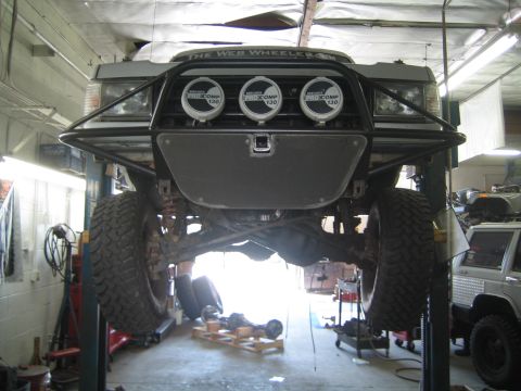 1990 Ford Bronco - Front view of a lifted truck raised on a shop lift, showing a bumper with three driving lights and a skid plate.