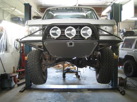 1990 Ford Bronco - lifted truck on a two-post shop lift displaying a tube front bumper with three round Pro Comp driving lights.