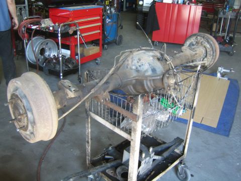 1990 Ford Bronco - removed rusty solid axle with a drum-brake backing plate sits on a wire shop cart amid red toolboxes in a garage.