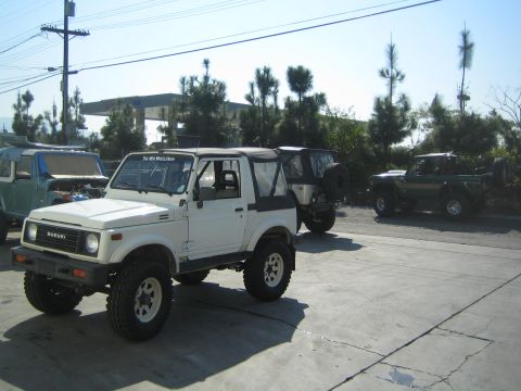 1990 Ford Bronco - white lifted Suzuki Samurai on off-road tires sits in a parking lot among other 4x4 vehicles and trees.