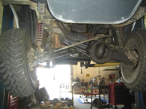 1990 Ford Bronco - Undercarriage of a lifted 4x4 on a shop lift showing coil springs, the front axle and steering linkage above the garage floor.