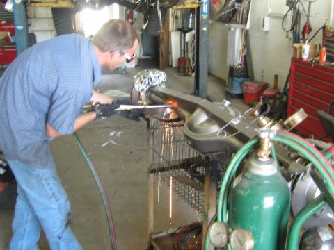 1990 Ford Bronco - man in a blue shirt uses an oxy-acetylene torch on an axle housing clamped to a cart, with sparks flying in a garage.