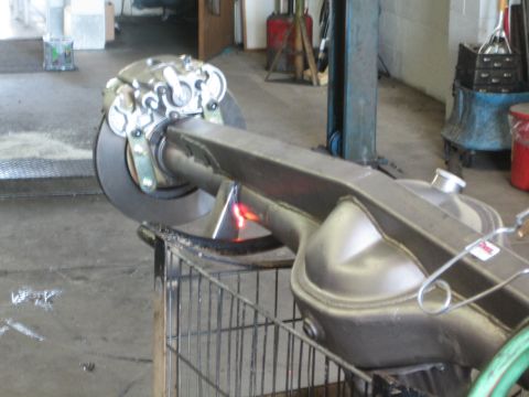 1990 Ford Bronco - axle housing with a disc-brake hub sits on a cart while a torch flame heats the tube inside a garage.