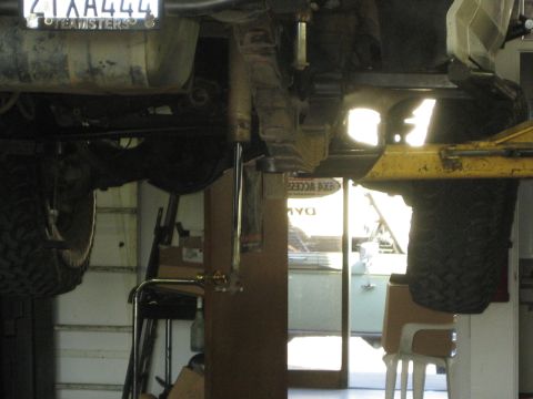 1990 Ford Bronco - Looking between two raised vehicles in a shop, an undercarriage with a Teamsters-frame license plate and off-road tires is visible.