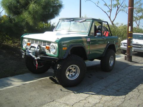 1990 Ford Bronco - lifted green early Ford Bronco with a white hood and front winch sits on a driveway riding on large off-road tires.