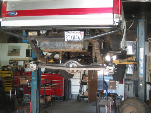 1990 Ford Bronco - underside of a red-and-white Ford truck raised on a shop lift, showing the exhaust, rear axle and a California plate reading 2TXA444.