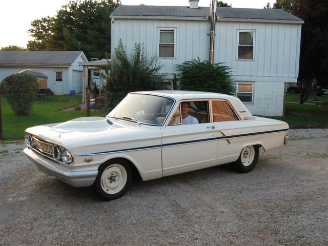 White 1964 Ford Fairlane two-door sedan on a gravel driveway by a white house