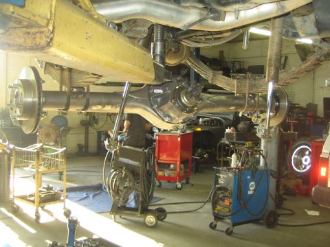 1990 Ford Bronco - vehicle raised on a lift reveals its driveshaft, differential and axle, with welders and tool carts in the busy shop below.