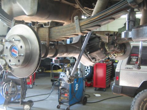 1990 Ford Bronco - underside view of a solid rear axle on leaf springs with a bare brake rotor, photographed on a lift in a repair shop.