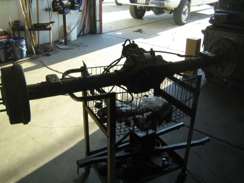 1990 Ford Bronco - complete rear axle assembly with brake backing plates sits on a wire shop cart in a garage, a lifted truck visible in the doorway.