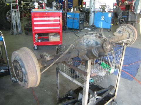 1990 Ford Bronco - rusty rear axle assembly with a brake drum rests on a wire cart in a welding shop with a red tool cabinet behind it.