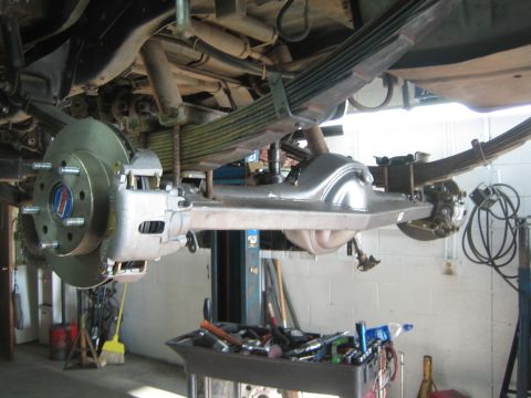 1990 Ford Bronco - freshly fabricated rear axle with a bare brake rotor and leaf springs is mounted under a vehicle raised on a two-post lift.
