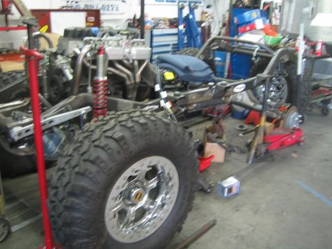 1990 Ford Bronco - bare tube-frame 4x4 chassis with a red coilover shock and an oversized mud tire sits mid-build in a fabrication shop.