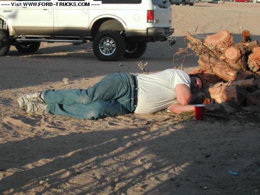 Man sleeping on sand near firewood pile, rear of white lifted Ford SUV in background