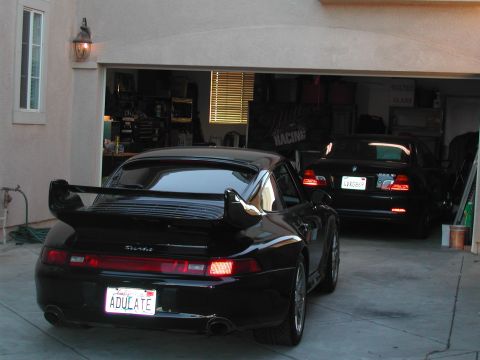 1997 Porsche 911 - A black Porsche 911 with a prominent rear wing is parked in front of a garage, with a black BMW visible inside.