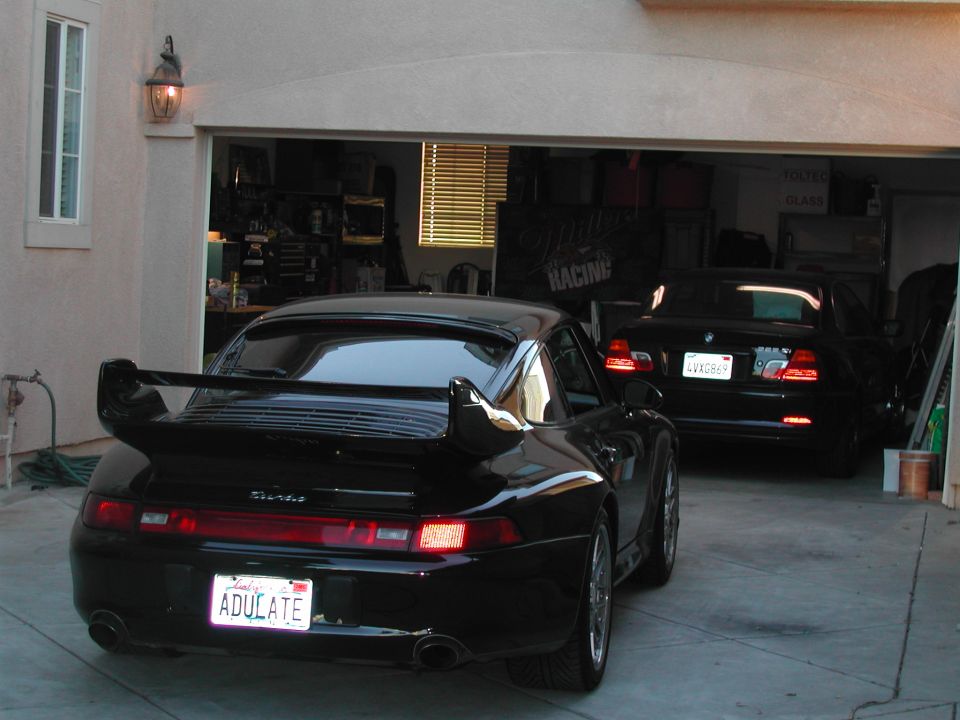 Black Porsche 911 Turbo rear view with biplane wing in driveway, black BMW in garage