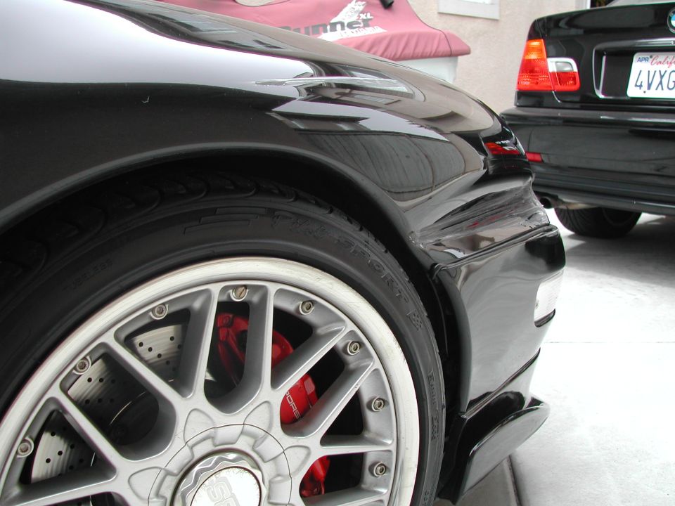 Black sports car rear wheel with silver multi-spoke rim and red calipers, black BMW behind