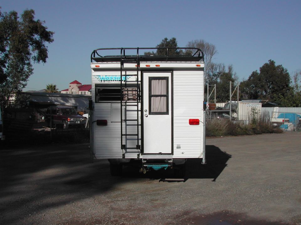 Rear view of a white truck camper with a ladder and roof rack. Another vehicle is visible in the background