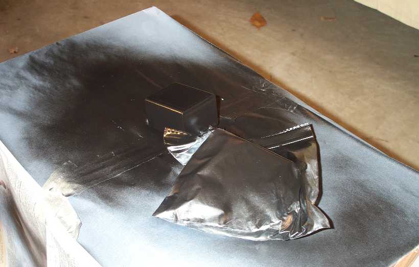 Small black-painted plastic box on black paper beside a plastic bag on a bench