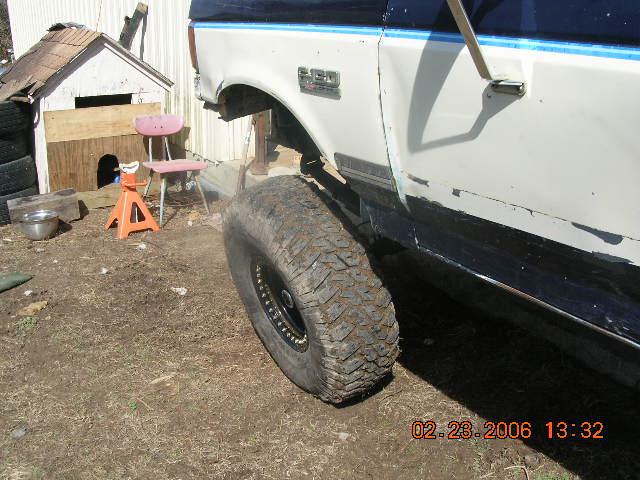 Blue and white F-150 front fender with mud tire removed and truck on a wood block