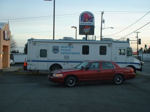 1999 Ford Crown Victoria - A red sedan is parked next to a Seattle Police Department van, with fast-food signs in the background.