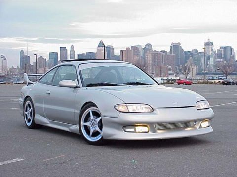 1994 Ford Probe - Silver coupe with a rear spoiler and custom wheels is parked in a lot with a city skyline behind it.