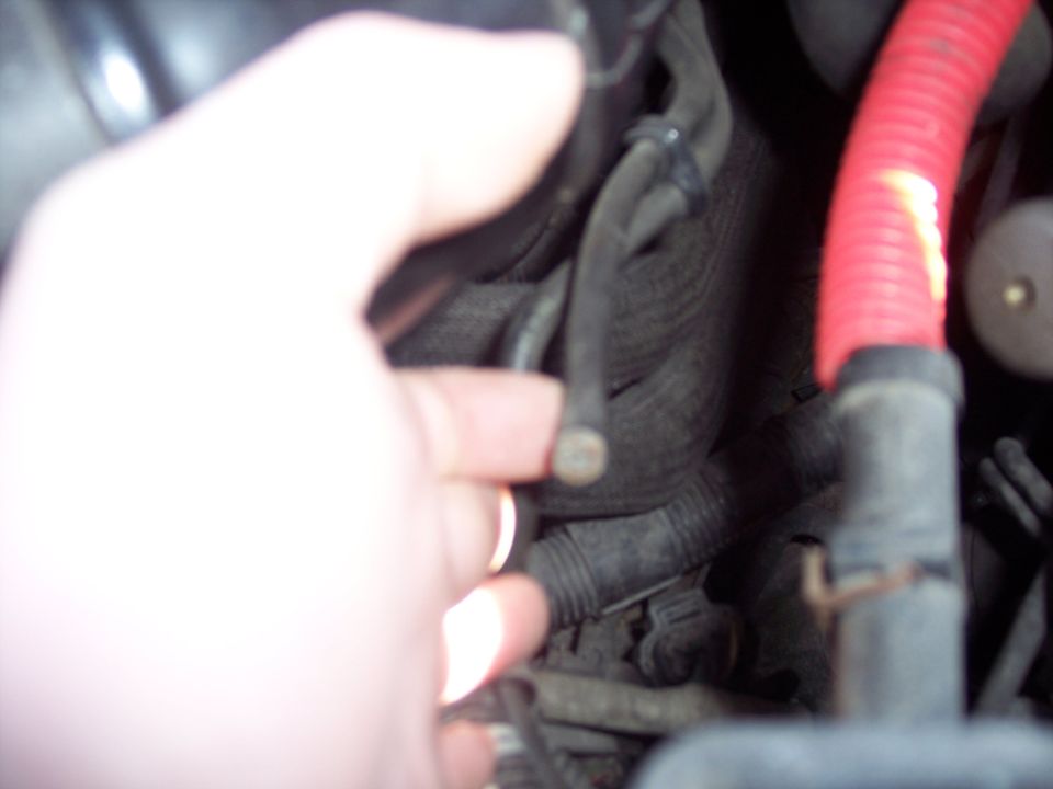 Hand pointing at a vacuum line in a dark engine bay wiring harness