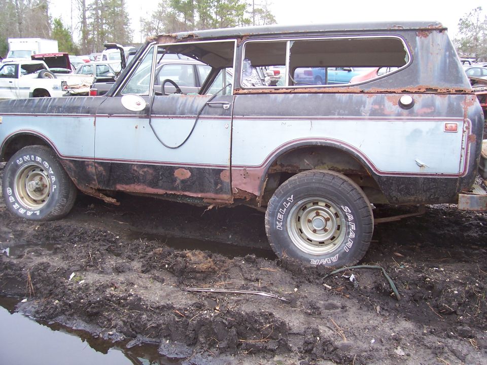 Rusty International Scout II parked in mud, side view. Other vehicles in background