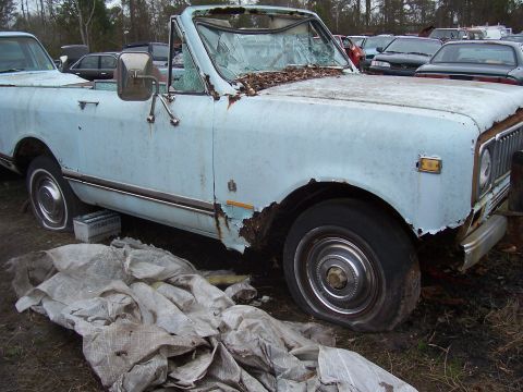 1978 Ford Bronco - A dilapidated light blue convertible SUV sits in a junkyard, showing severe rust and damage.