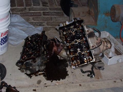 1996 Ford Probe - Two engine cylinder heads, covered in oil, lie on a concrete floor next to a bucket.