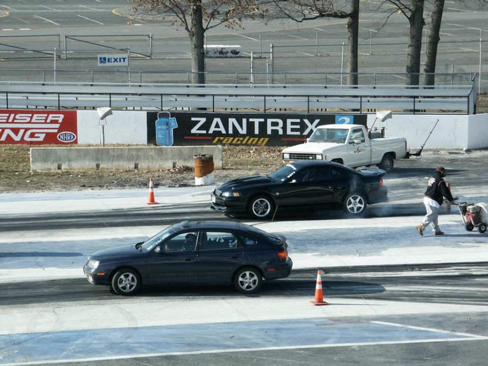 Dark sedan and black coupe racing on a drag strip with a white pickup truck in the background