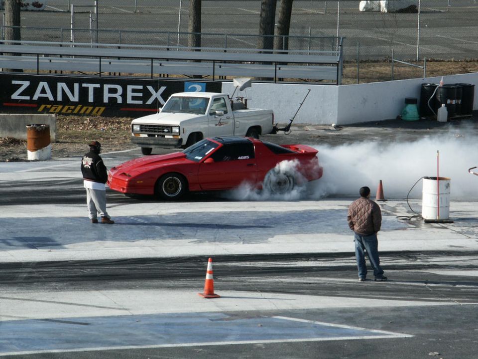 Red sports car doing a burnout on a drag strip with a white pickup truck in the background