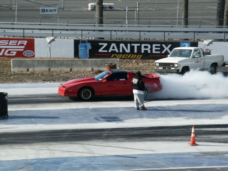 Red Pontiac Firebird doing a burnout next to a white pickup truck at a drag strip