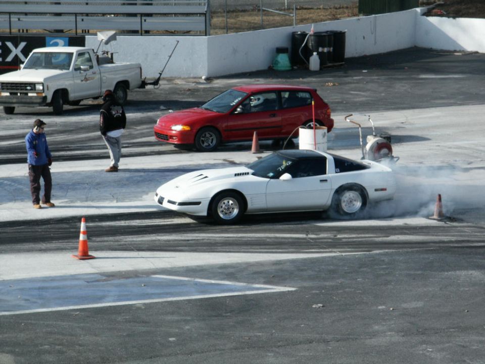 White Corvette C4 doing a burnout on a drag strip with a red hatchback and a pickup truck in the background