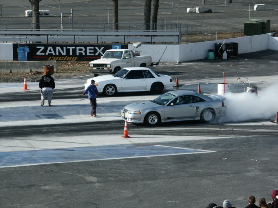 Silver Ford Mustang doing a burnout on a drag strip, white car and truck in background