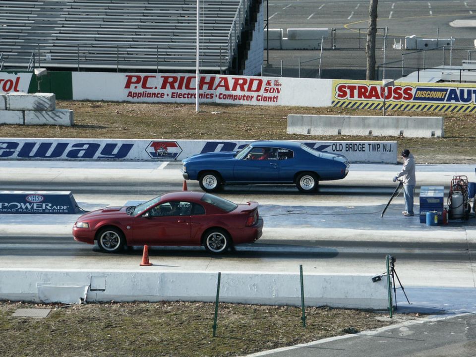 Red coupe and blue sedan racing down a drag strip with grandstands in the background