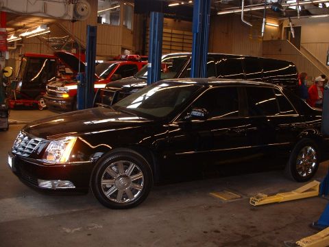 1999 Ford Crown Victoria - A black Cadillac sedan is parked in a garage with other vehicles, including red trucks. The SUPERMOTORS.NET logo is visible.