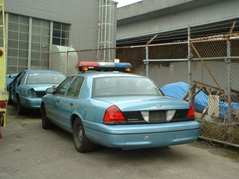 1999 Ford Crown Victoria - Two light blue police cars are parked in an outdoor lot, one behind the other.